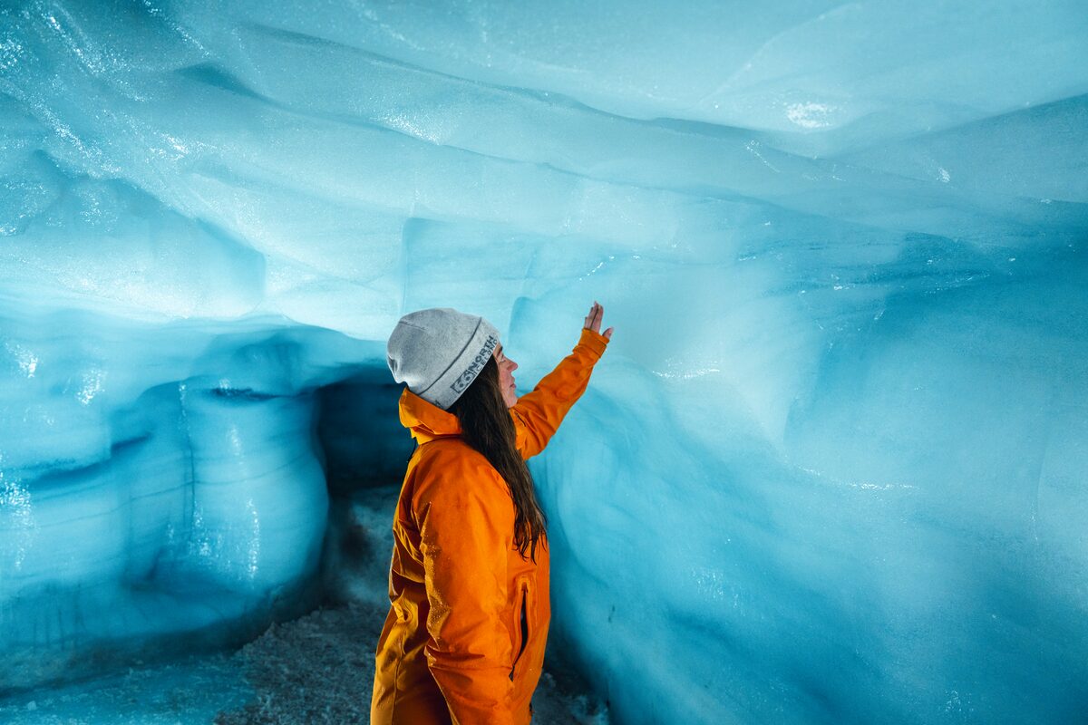 Woman in orange coat and white beanie hat touching blue ice tunnel walls at Langjokull