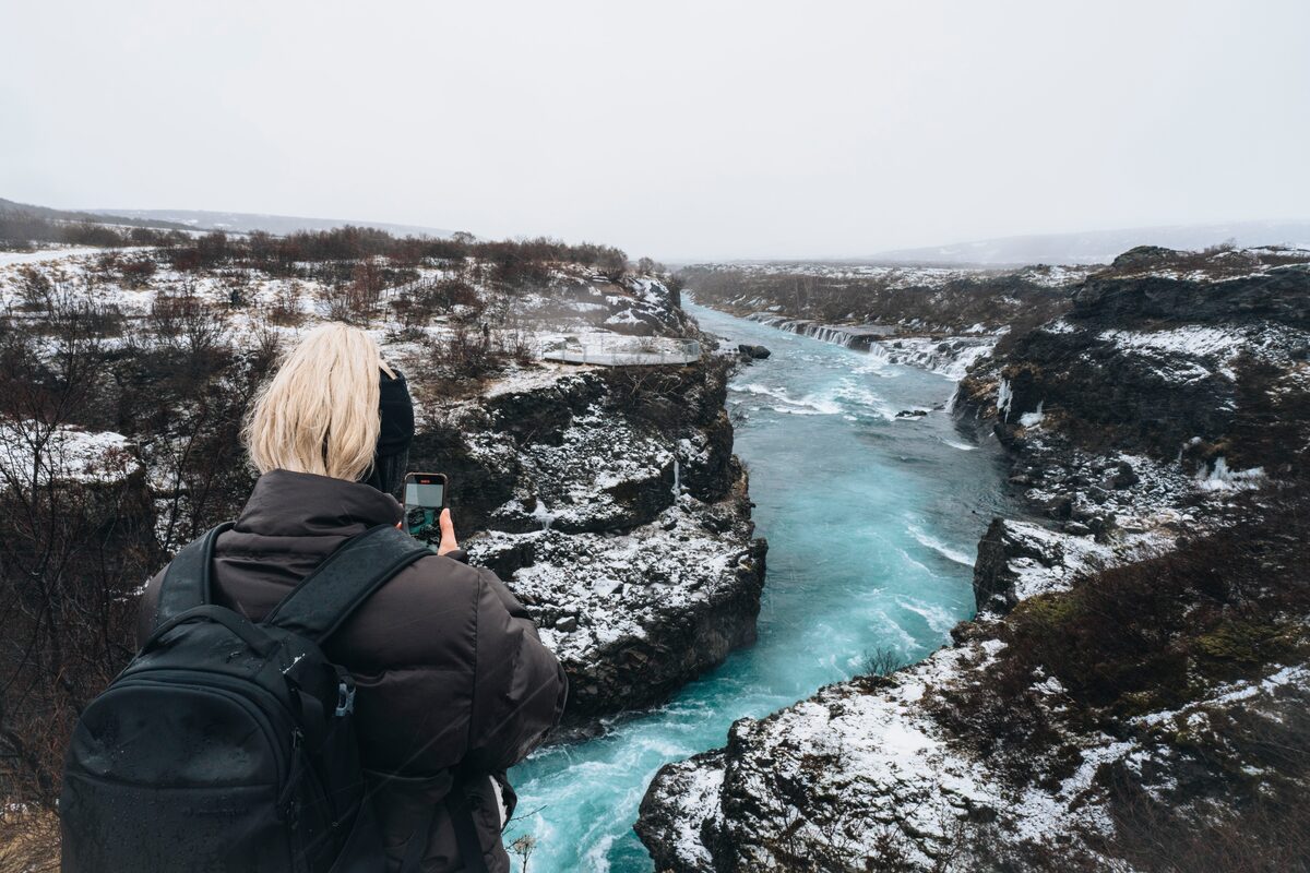 A lady dressed in a black jacket and backpack takes a photo of Barnafoss from the top.