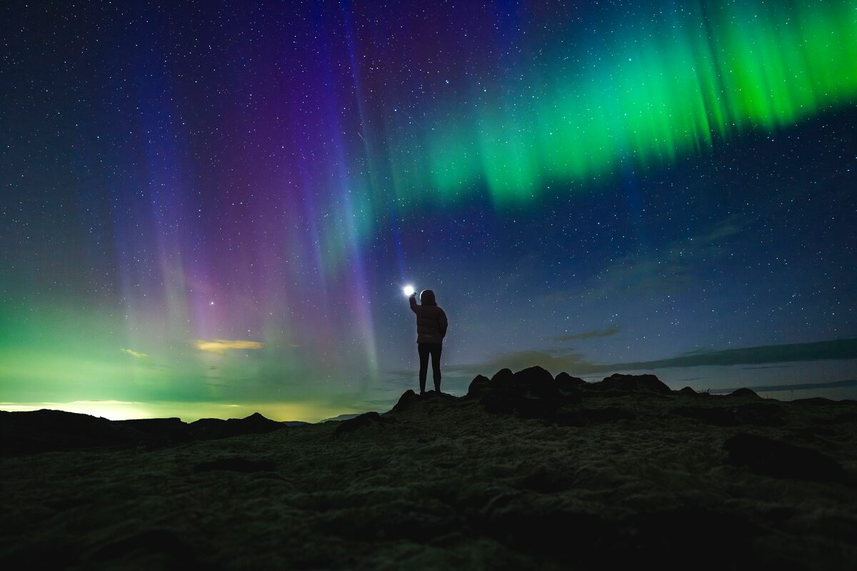 Silhouette of person standing in night sky full of northern lights holding phone light up