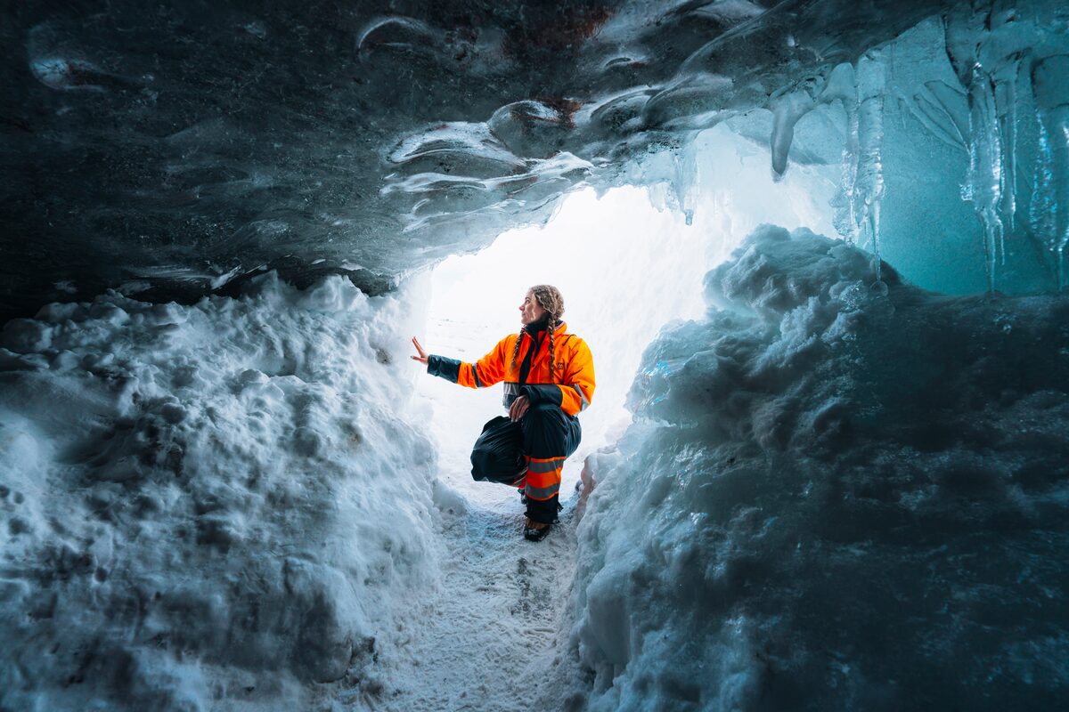 Young female wearing orange snowmobiling suit crouching inside ice cave entrance