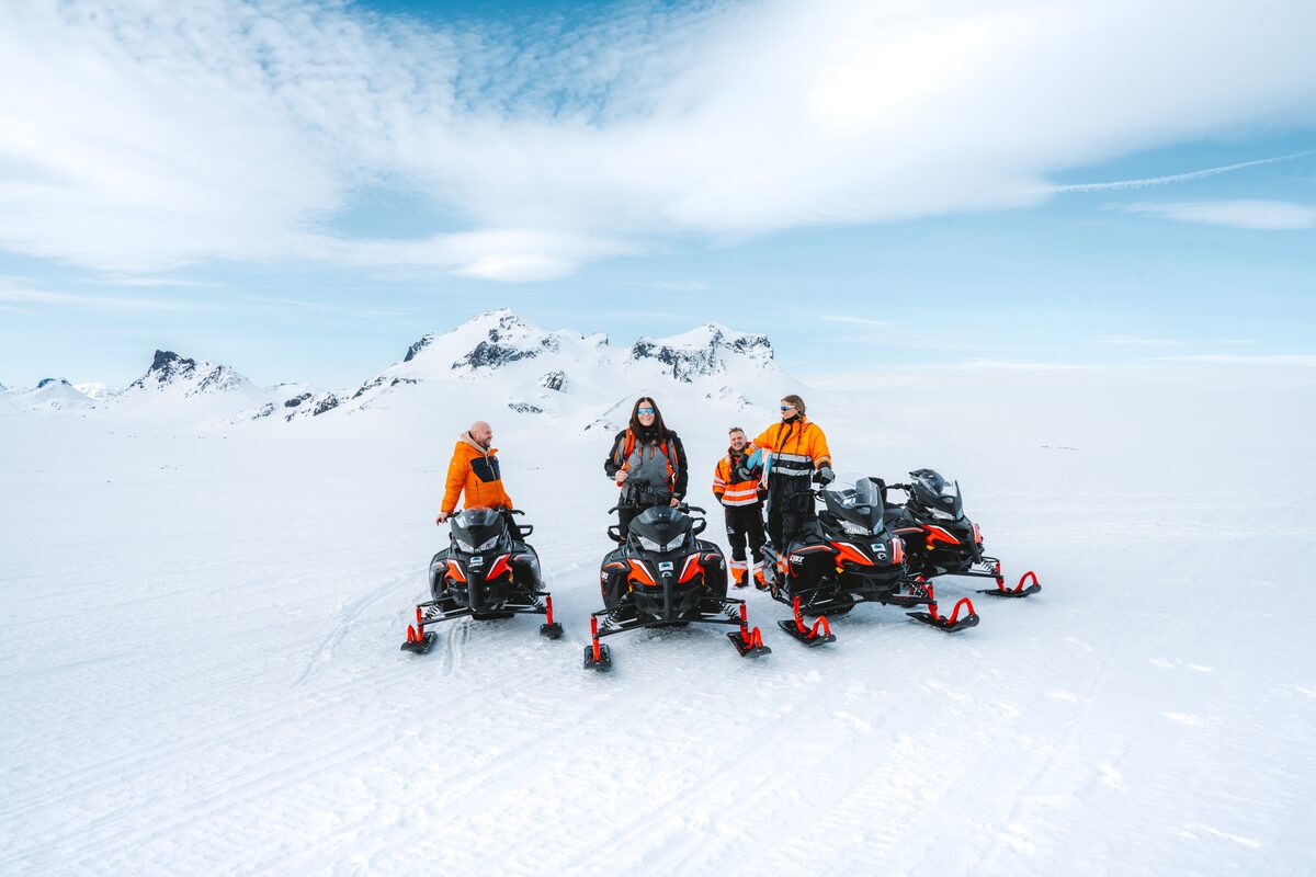 Small group of people on snowmobiles in front of huge Langjokull glacier with blue skies.