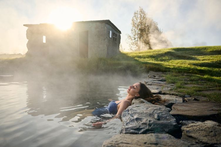 Lady bathing in secret lagoon at sun down next to old Icelandic stone building.