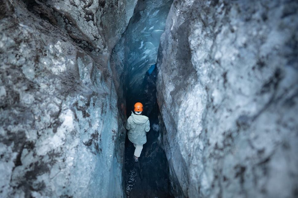 Back of person with orange safety helmet walking into ice cave entrance, beautiful crystal wall patterns from ice.