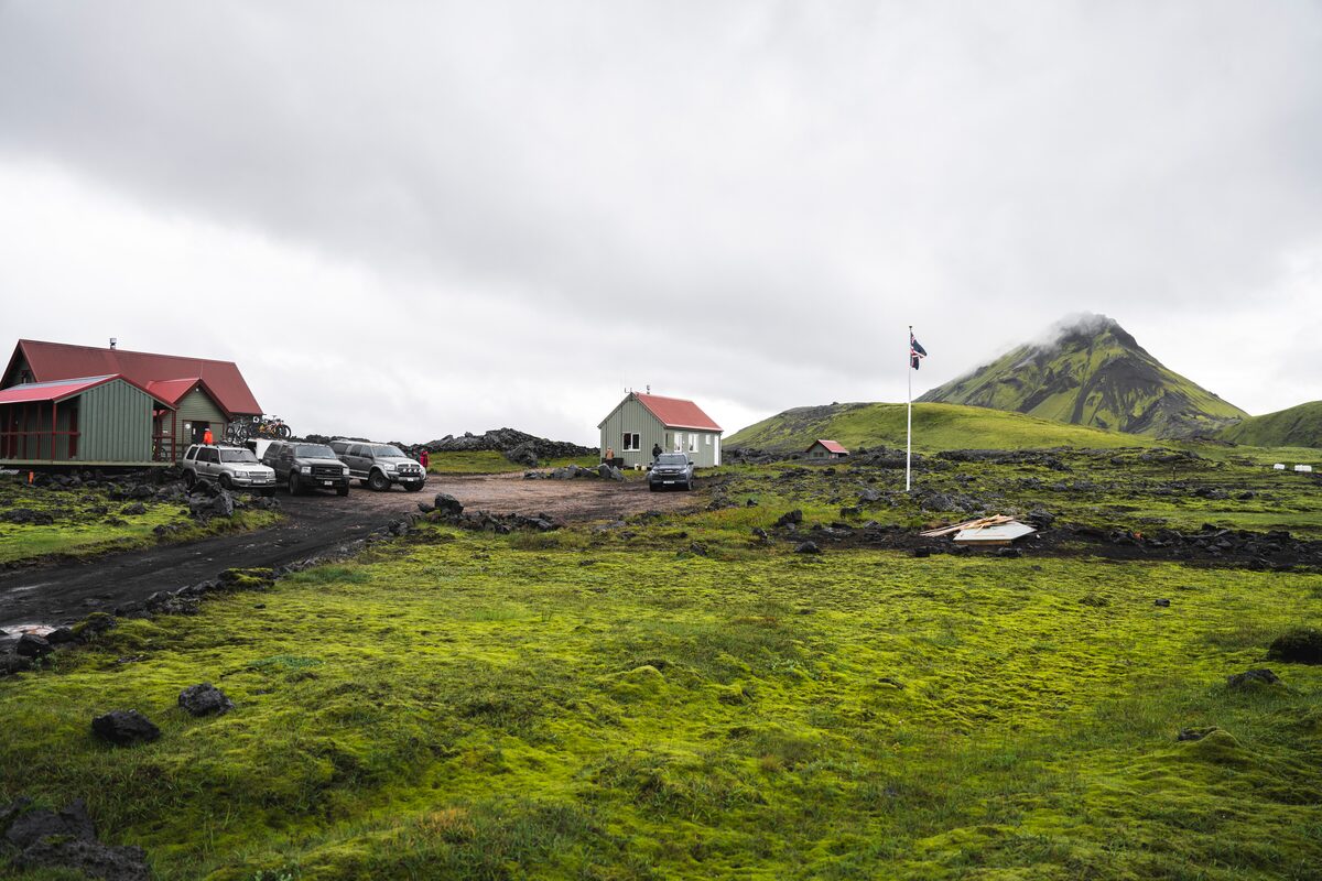 Cars parked at Hvannigil base huts in scenic moss covered hiking area with Icelandic flag.