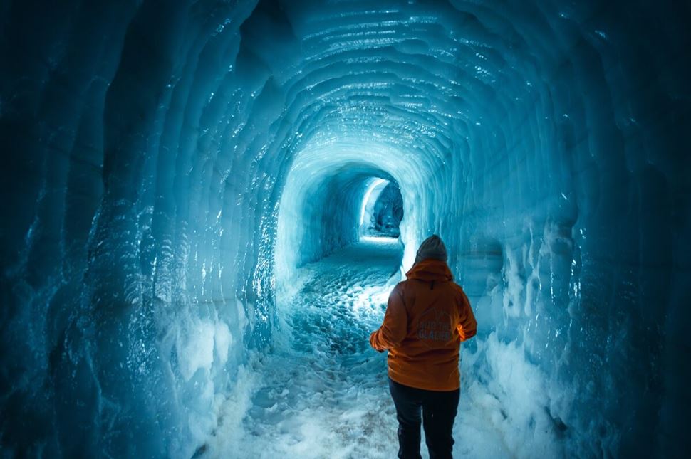 Woman walking through blue walled Langjokull ice tunnel with light reflecting on ice wall.