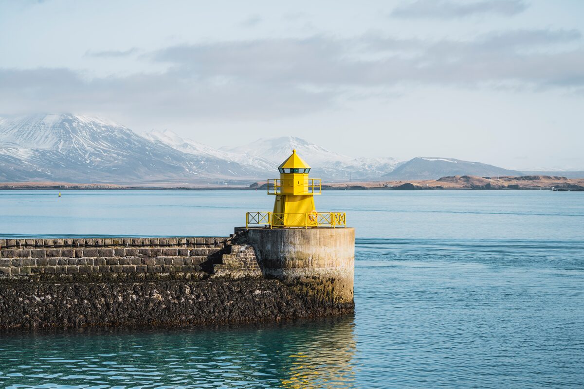 Small yellow lighthouse at Reykjavik harbor, with view of snowy mountains in distance.