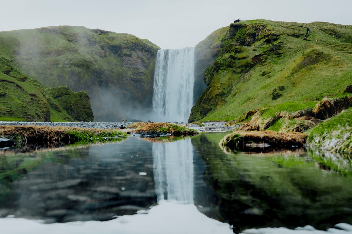 Skogafoss waterfall and reflection into nearby river surrounded by green hills in summer.