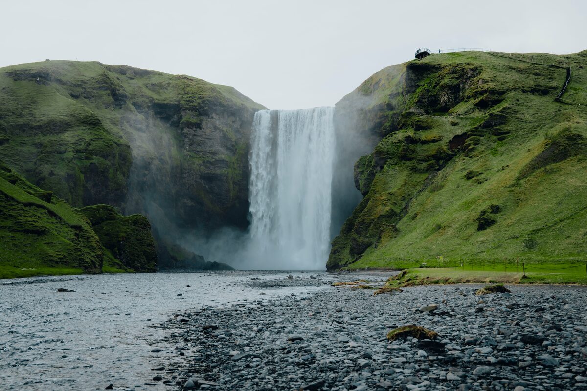 Skogafoss waterfall flowing into river with black pebble covered ground and surrounding green hills during summer months.