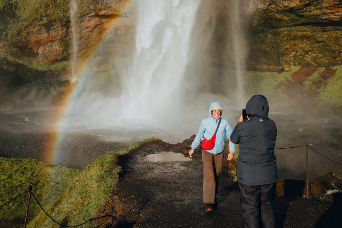 Two happy tourists psoing for a photo underneath Seljalandsfoss waterfall by rainbow in south Iceland.