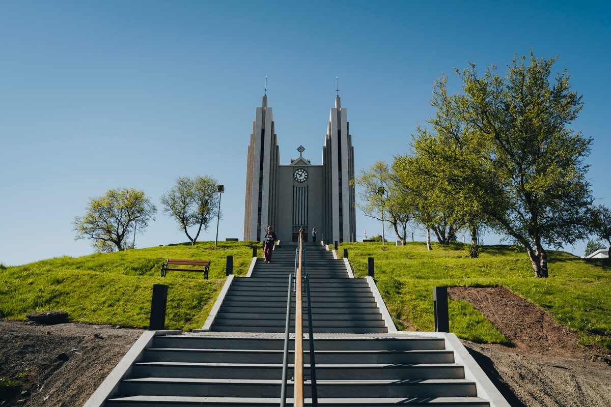 Steps leading up to Akureyri church, Akureyrarkirkja on a grass covered hill and surrounding trees in sunshine.