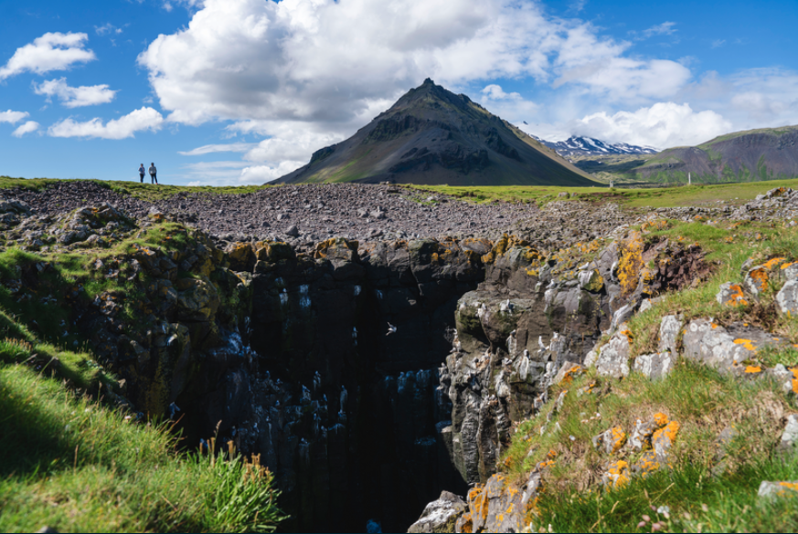 Two tourists standing in front of Arnarstapi mountain by grass covered cliff area in summer.