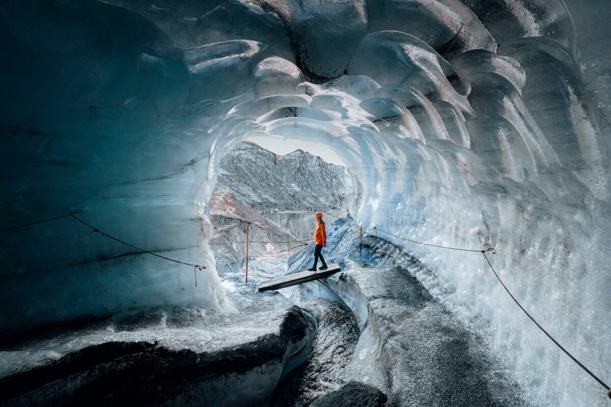 Woman walking on a wooden plank bridge inside Katla ice cave, surrounded by shimmering blue and black ice walls.
