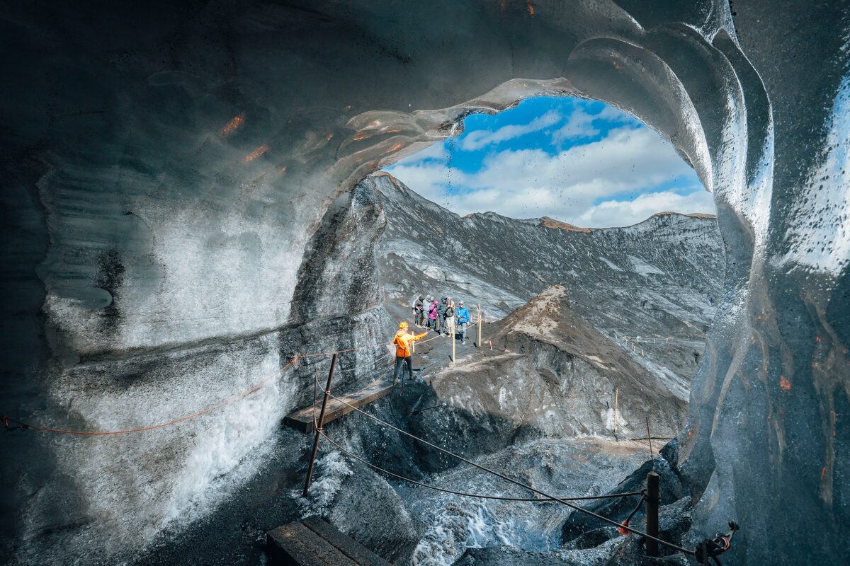 Tourists walking near the entrance of Katla ice cave, surrounded by dark volcanic terrain and glacial ice formations.