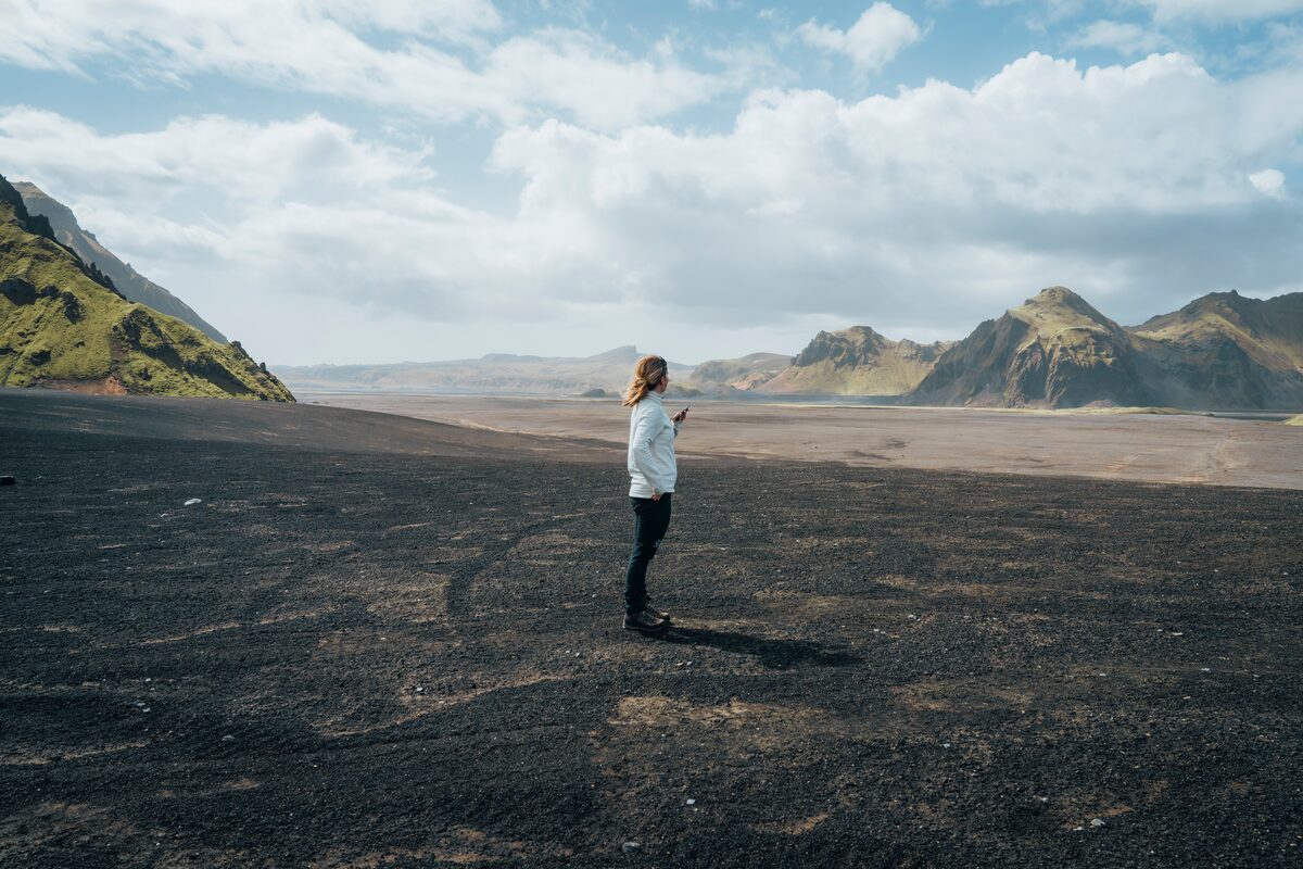 Woman posing for a photo on volcanic ash fields between Katla volcano and a moss-covered mountain, with contrasting dark and green landscapes in the background.