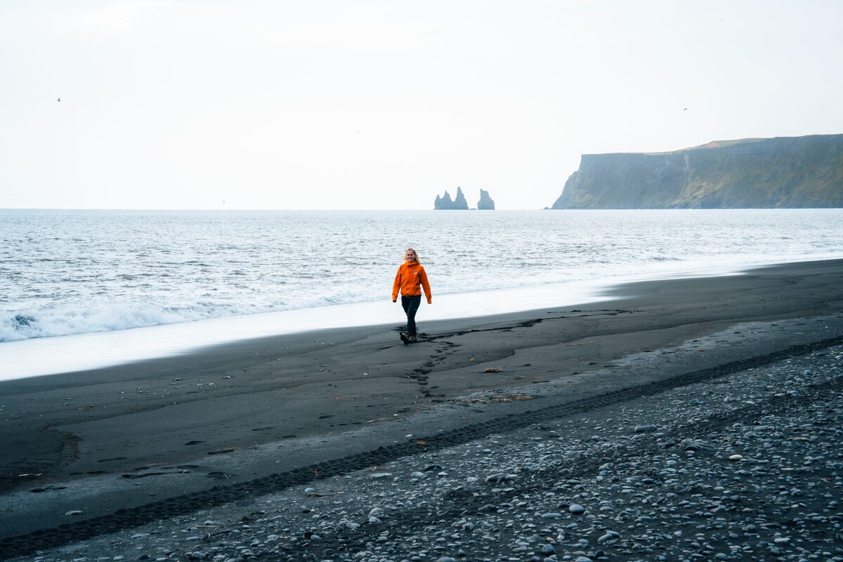 Woman walking along Reynisfjara black sand beach on Iceland’s South Coast under an overcast sky, with basalt sea stacks in the distance.