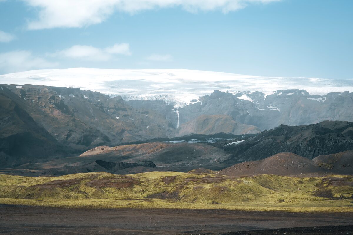 Volcanic mountain landscape with patches of moss-covered ground and the Mýrdalsjökull glacier visible in the background under a partly cloudy sky.