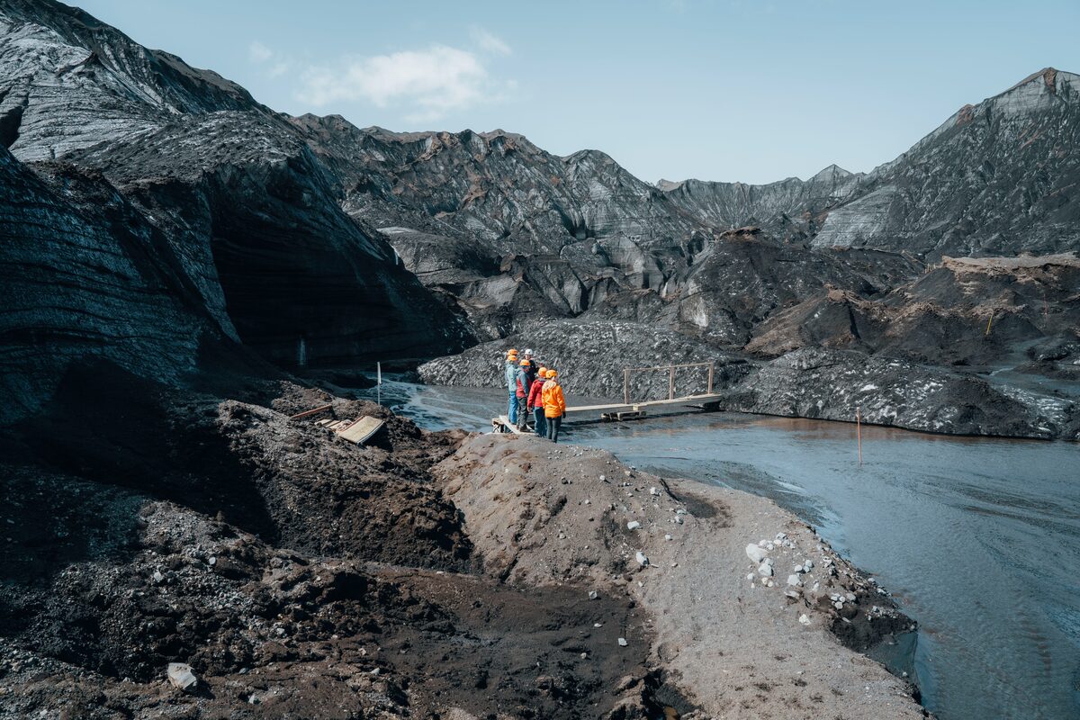 People standing near a wooden plank bridge over a stream in a rugged volcanic mountain area, surrounded by dark volcanic terrain and steep slopes.