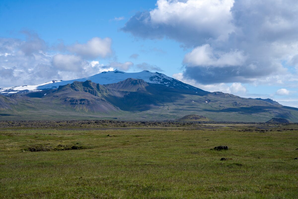 Panoramic view of Snæfellsjökull glacier in Iceland, with its snow-capped peak rising above rugged volcanic terrain, surrounded by green fields.