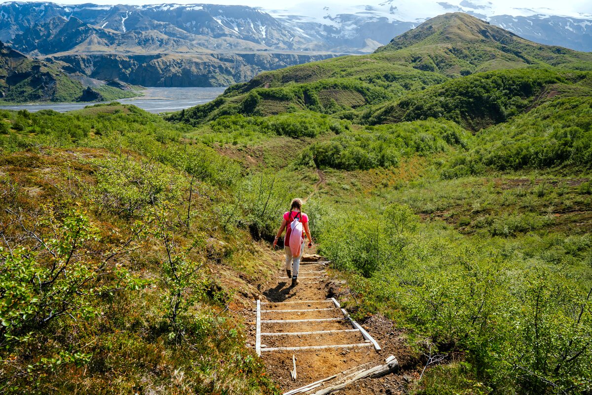 Female hiker walking along a scenic trail in Þórsmörk, Iceland, surrounded by green summer vegetation, dramatic mountain peaks, and a clear blue sky.
