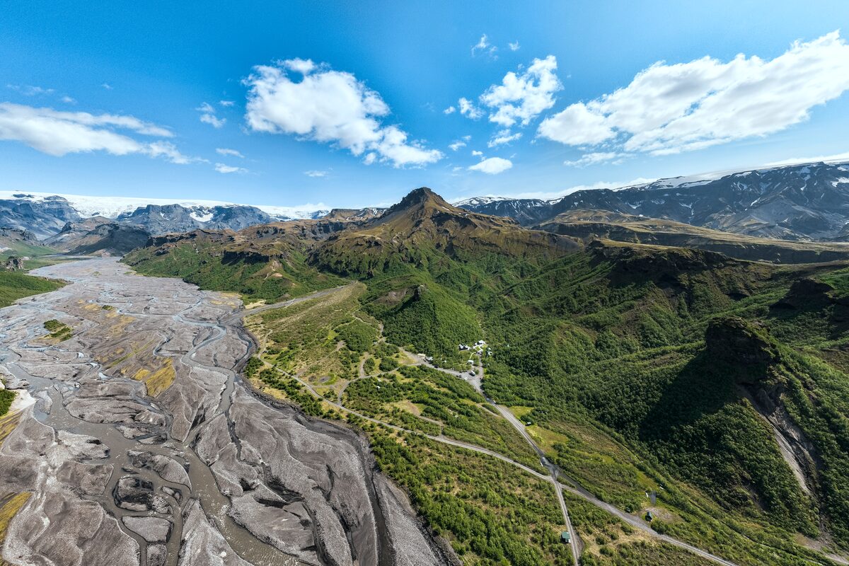 Aerial view of Þórsmörk Valley in summer, showcasing a lush green landscape with winding rivers, rugged mountain ridges, and patches of birch forest beneath clear blue skies.