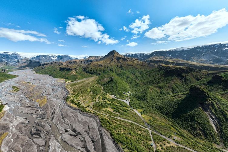 Aerial view of Þórsmörk Valley in summer, showcasing a lush green landscape with winding rivers, rugged mountain ridges, and patches of birch forest beneath clear blue skies.