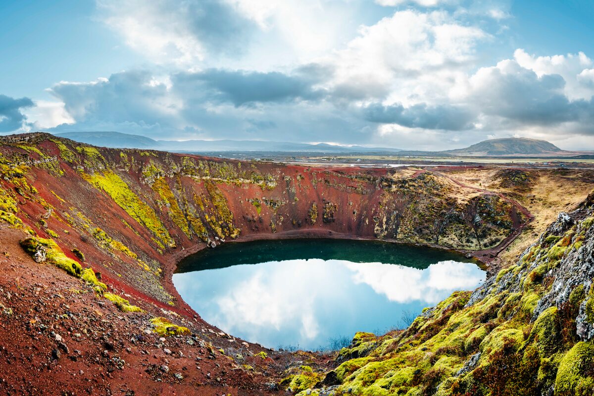 Moos covered Kerid volcanic crater edge and blue cloudy sky reflecting into crater lake.