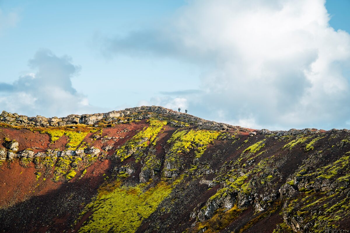 Two tourists walking round Kerid volcanic rock and moss covered crater edge, on a brright sunny day.