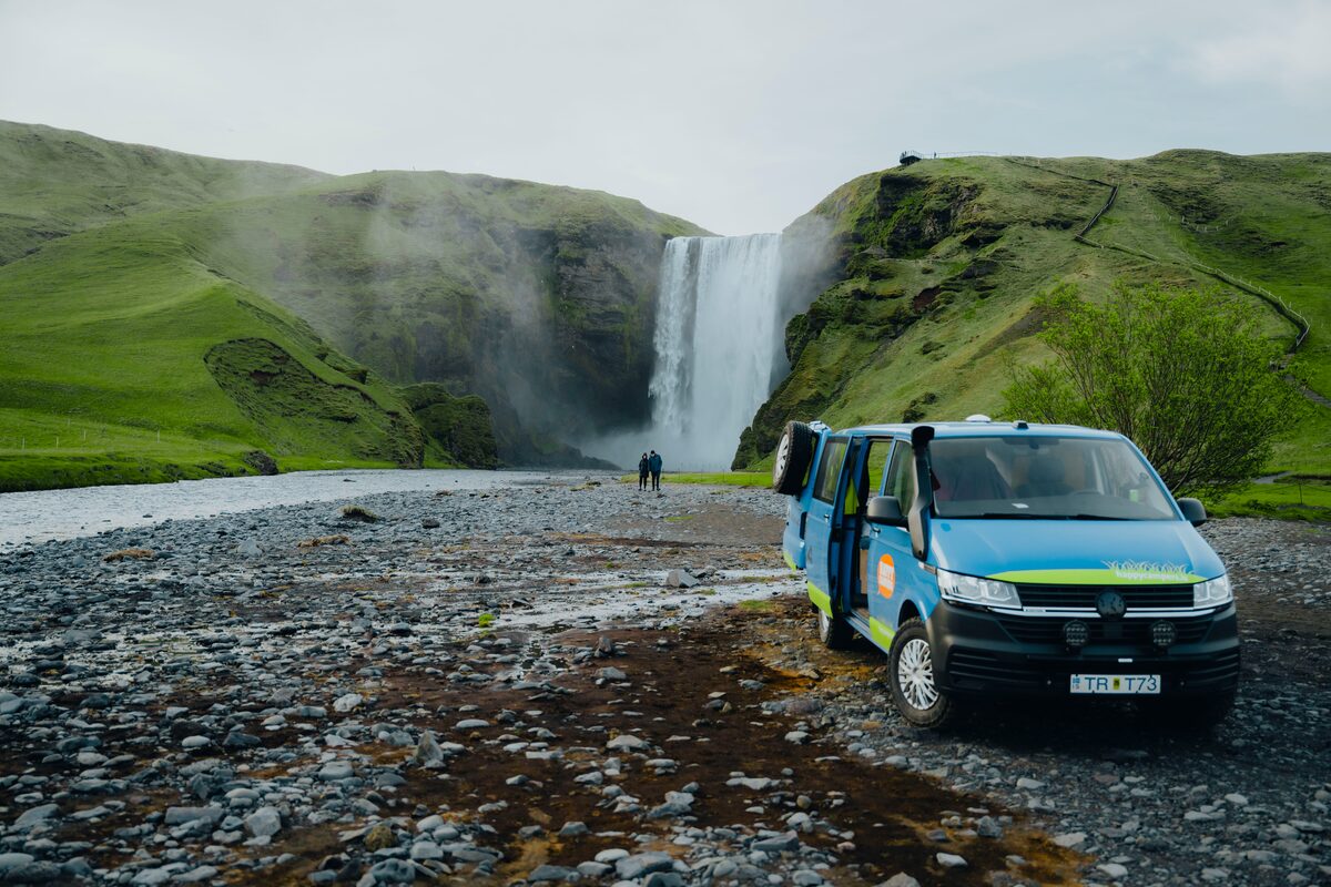Blue Happy Campers rental camping van parked outside Skogafoss waterfall in summer.
