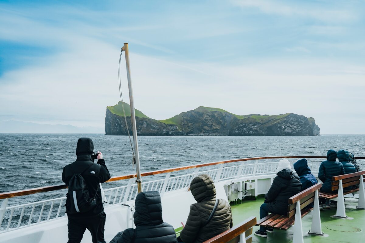Group of tourists travelling on a boat towards Westmann islands, photographing lonely house on Ellidaey island.