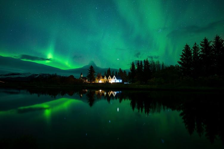 Houses at Thingvellir national park surrounded by trees during beautiful bright aurora lights.