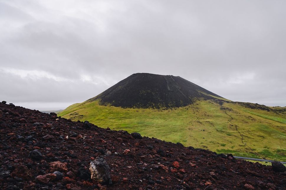 Top view of Eldfell Volcano under overcast grey skies, showing the rugged volcanic crater rim and dark lava flows spreading across the landscape below.