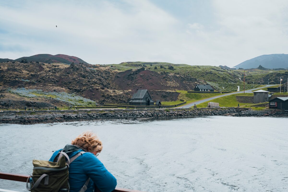 View from a ferry approaching the Vestmannaeyjar Islands, with the striking black Viking-style church visible near the shoreline. Surrounding the church is a sweeping grassy landscape, set against dramatic cliffs and a cloudy sky in the background.