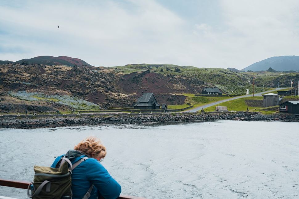 View from a ferry approaching the Vestmannaeyjar Islands, with the striking black Viking-style church visible near the shoreline. Surrounding the church is a sweeping grassy landscape, set against dramatic cliffs and a cloudy sky in the background.