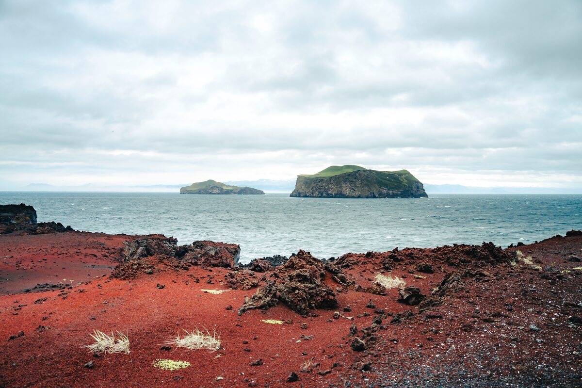 View of a rugged red rock foreground leading to the sea, with gentle waves meeting the shoreline. In the distance, Heimaey Island rises from the ocean, its cliffs and green slopes partially shrouded by low clouds. 