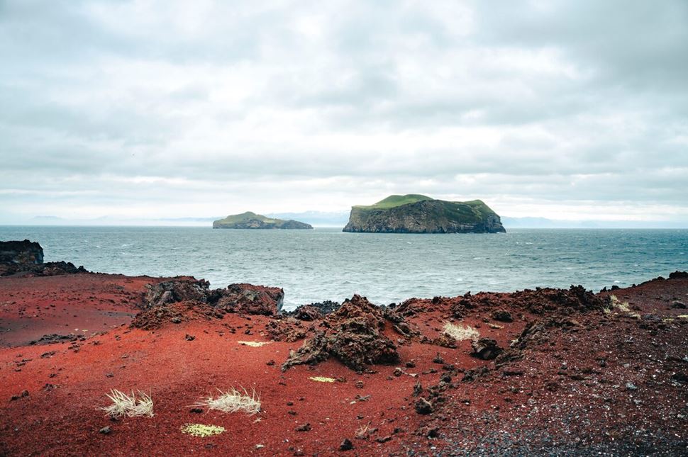 View of a rugged red rock foreground leading to the sea, with gentle waves meeting the shoreline. In the distance, Heimaey Island rises from the ocean, its cliffs and green slopes partially shrouded by low clouds.