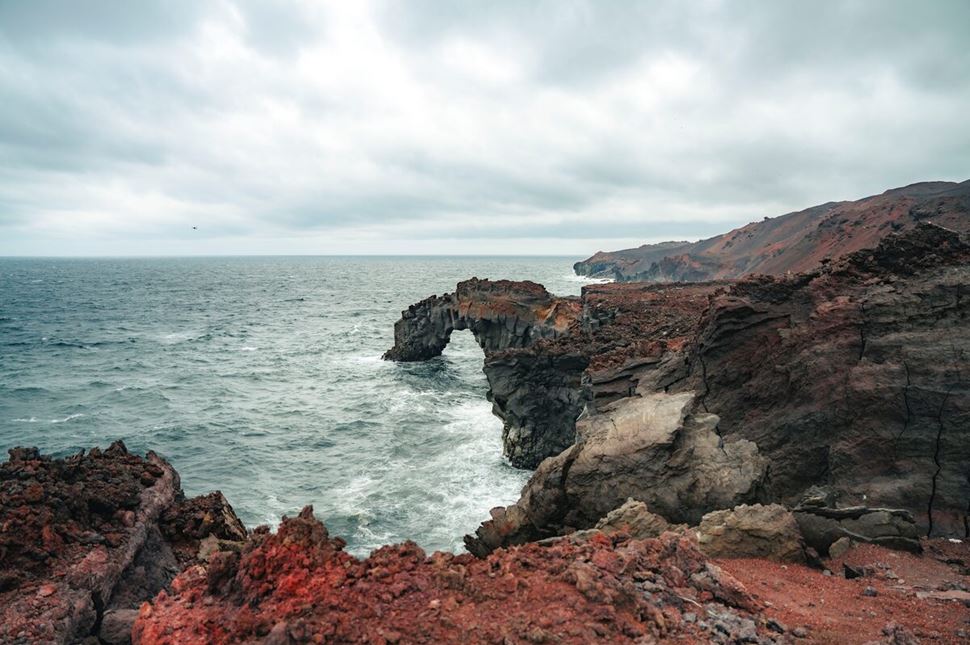 Coastal red rock terrain and arch on Vestmannaeyjar Island, with rugged cliffs and weathered formations stretching toward the sea.