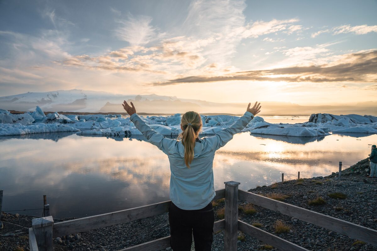 Lady posing with arms up in the air, standing viewing Jokulsarlon glacier lagoon during sunset.
