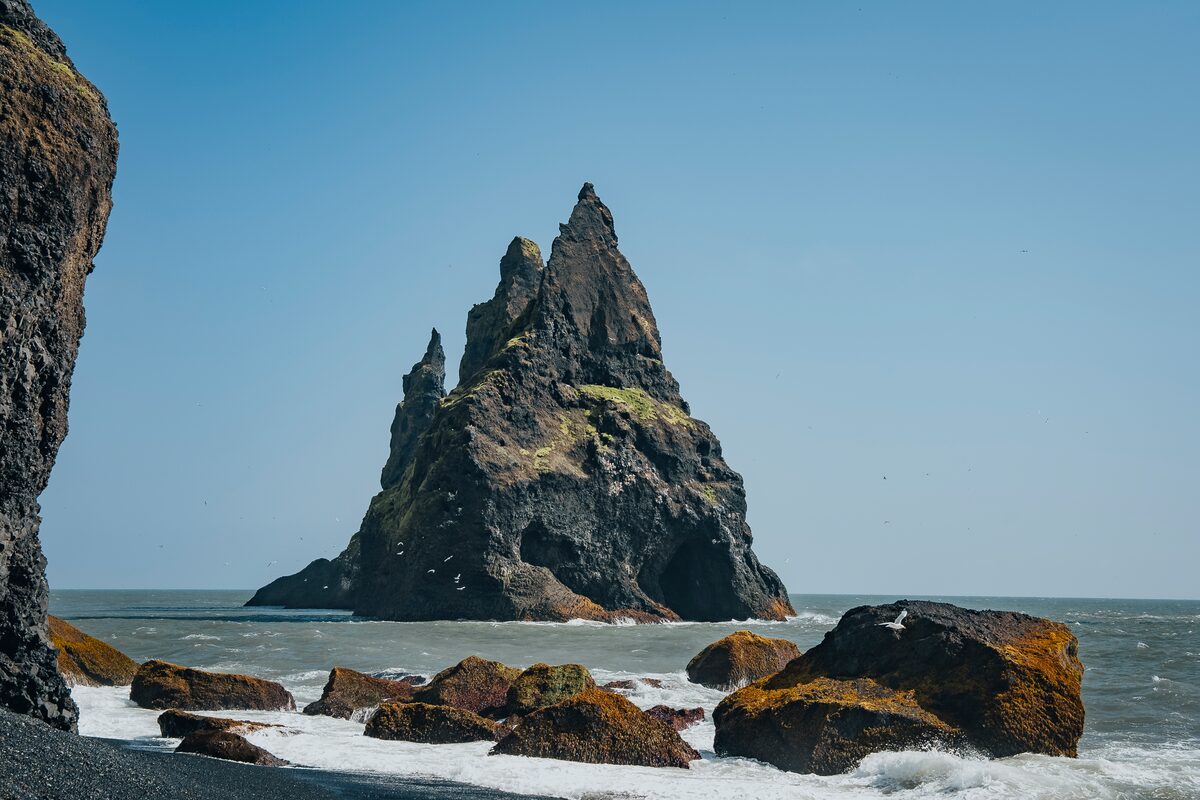 Reynisfjara Beach with a large sea stack rising from the ocean, accompanied by smaller rock formations near the cliffside under clear blue skies.