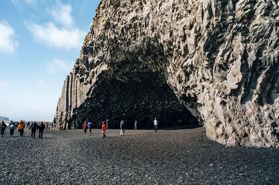 Tourists walking around Halsanefshellir Cave, exploring the rocky formations and dark cavern openings along the coastline.