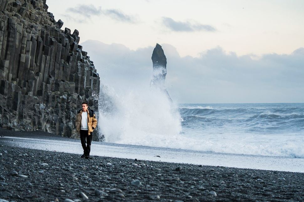 Harsh waves crashing onto the black sand of Reynisfjara Beach as a tourist poses for a photo near the shoreline.