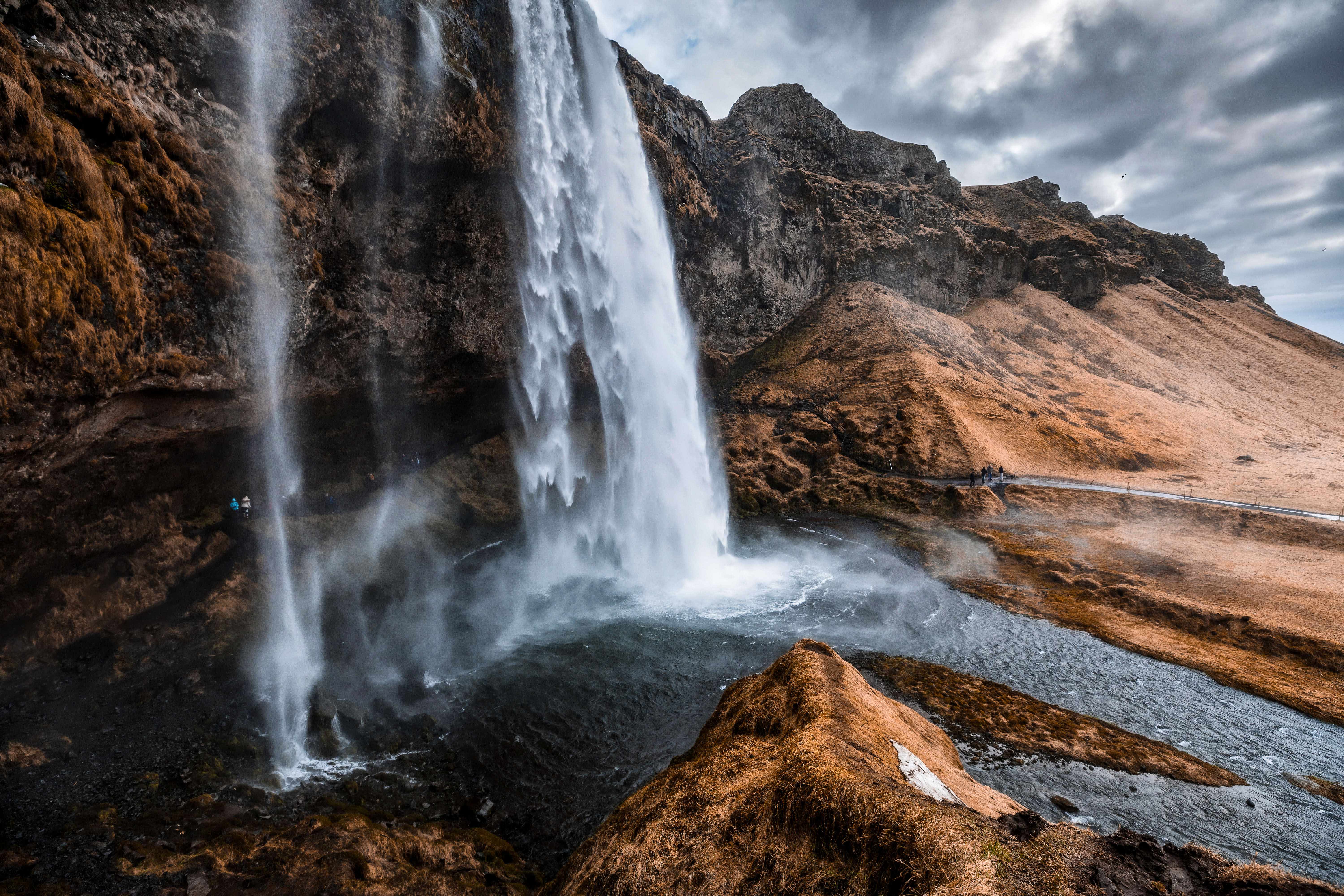 People walking behind Seljalndsfoss waterfall, autumnal landscape with snow starting to settle on ground.