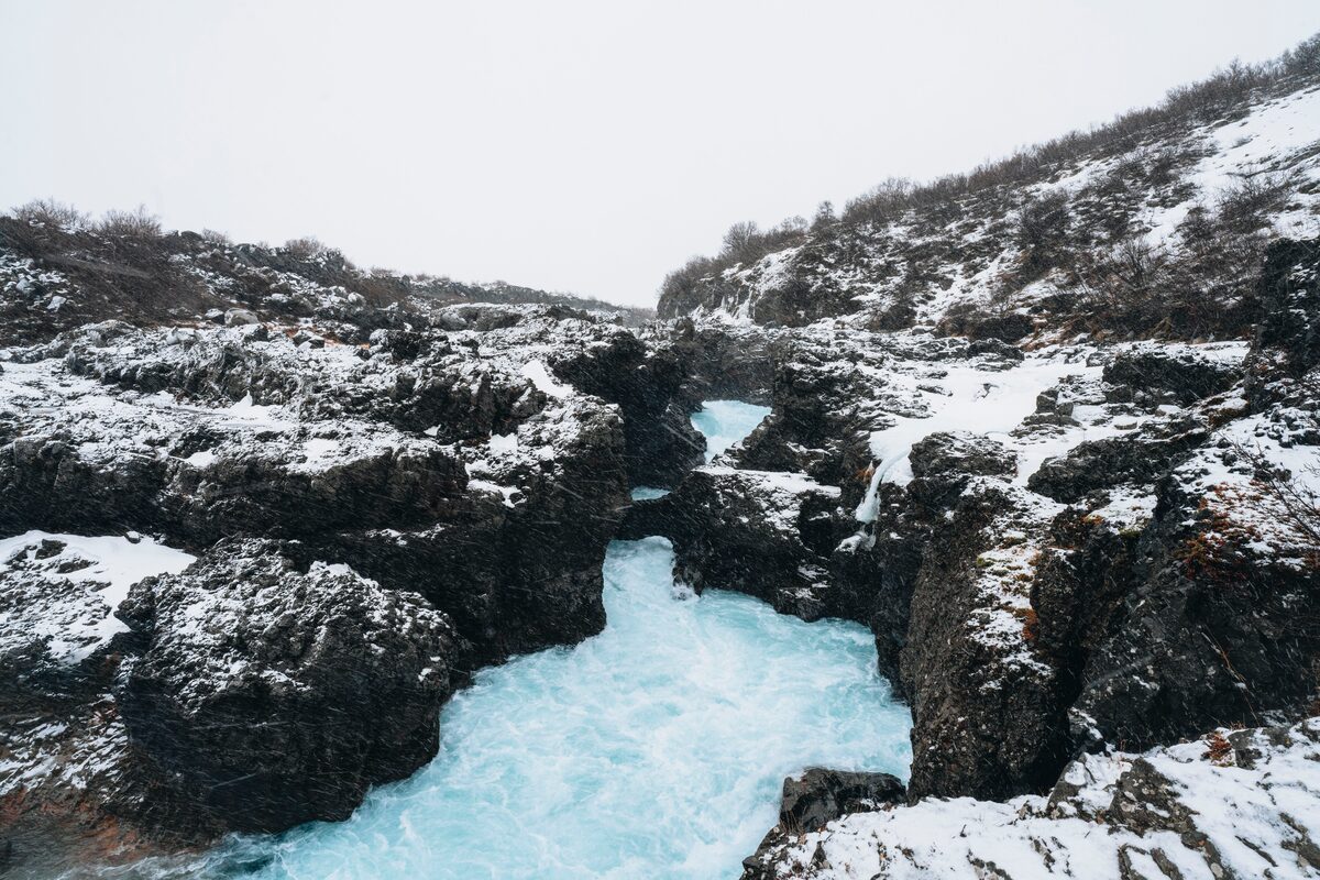 Snow covered cliffs at bright blue Barnafoss waterfall in Iceland during winter.