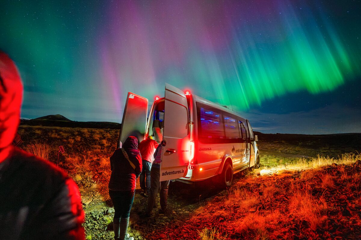 Tour group exiting white Arctic Adventures van underneath bright northern lights, red van lights reflecting onto grass.
