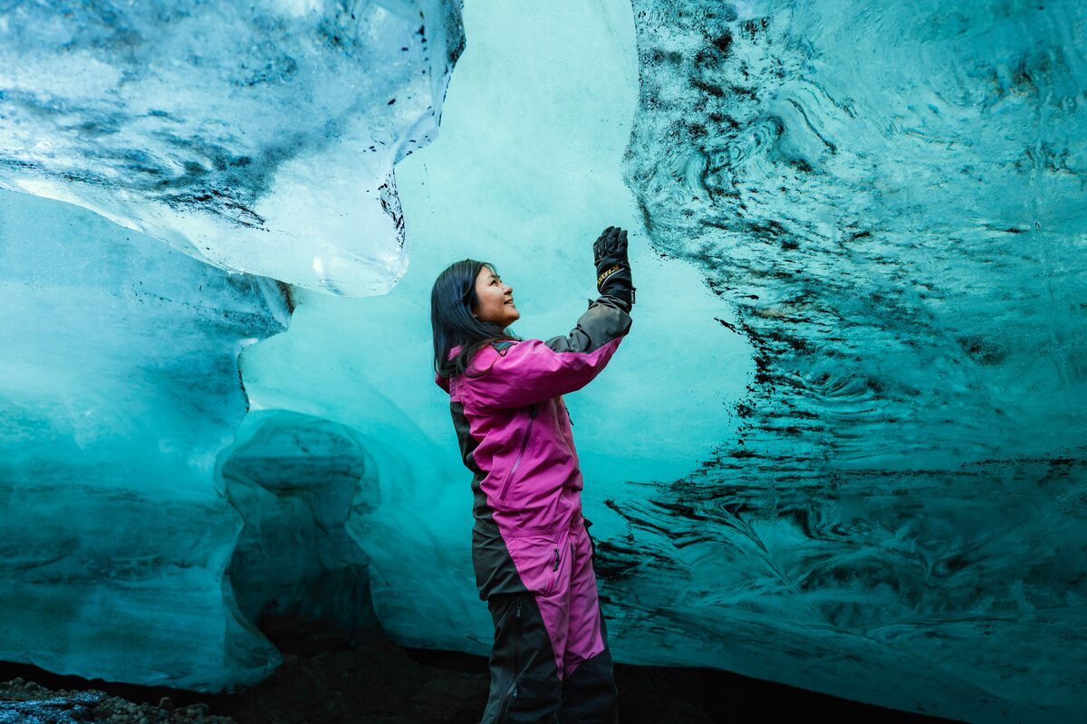 Female wearing pink snow suit, reaching to turquoise blue ice cave wall, surrounded by textured crystal walls at Langjokull.