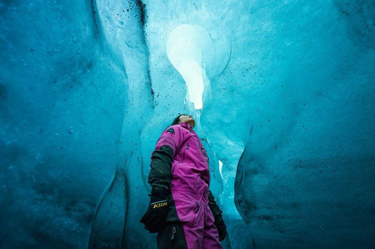 Female wearing pink snow looking up to ice cave roof, standing inside turquoise blue ice cave at Langjokjull glacier.