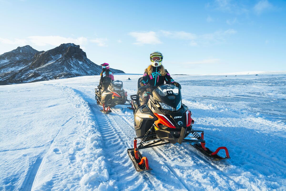 Duo snowmobiling down Langjokull glacier slopes in full snowmobiling protective gear.