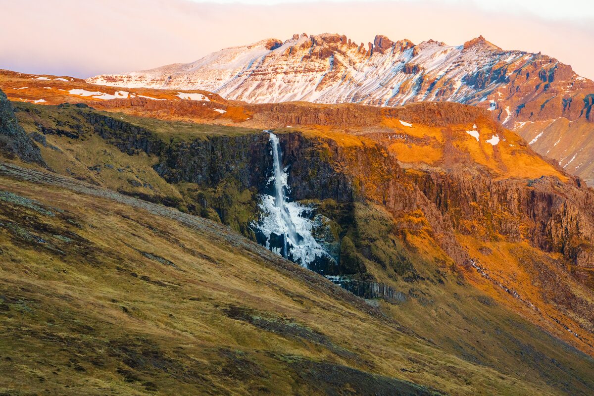 Crashing waterfall in Snaefellsnes Peninsula, snow covered Fjords in background during sunset.