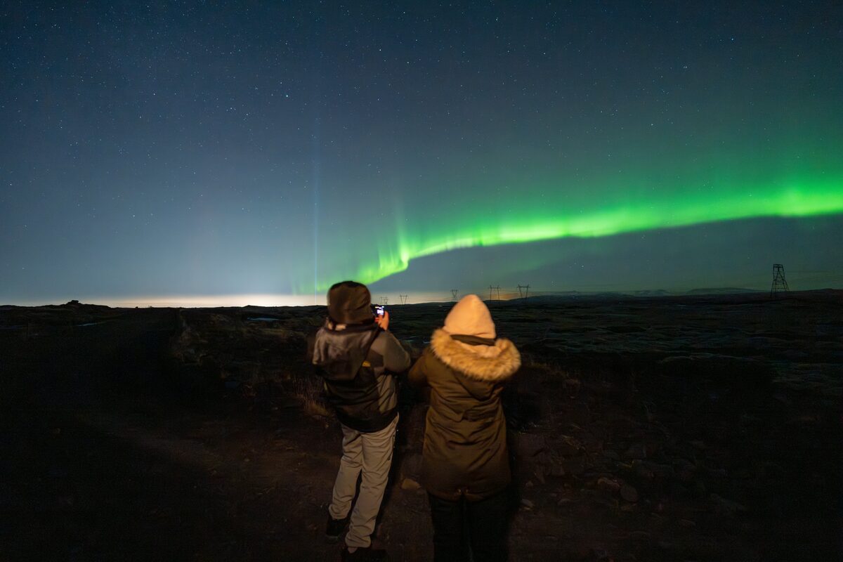 Couple standing together looking to night sky, capturing northern lights on phone camera.