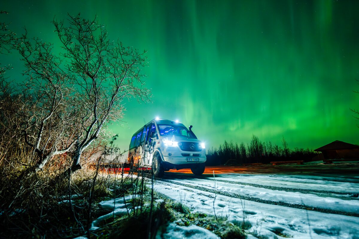 Arctic Adventures white tour bus with lights on in night, parked on snow under northern lights.