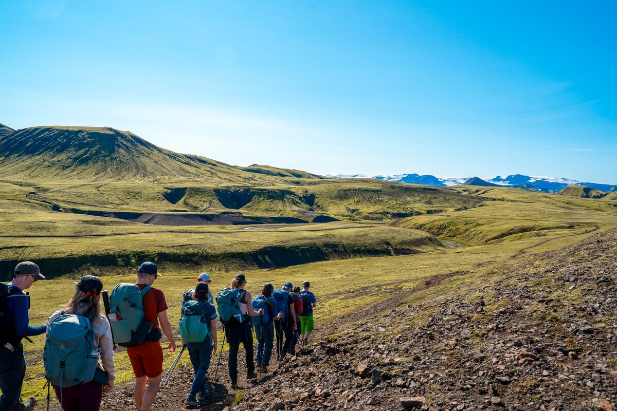 Small group hiking across moss covered terrain with views of moss covered mountain in distance and blue skies.